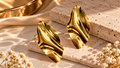 Gold-tone sculptural earrings with flowing folds displayed on travertine